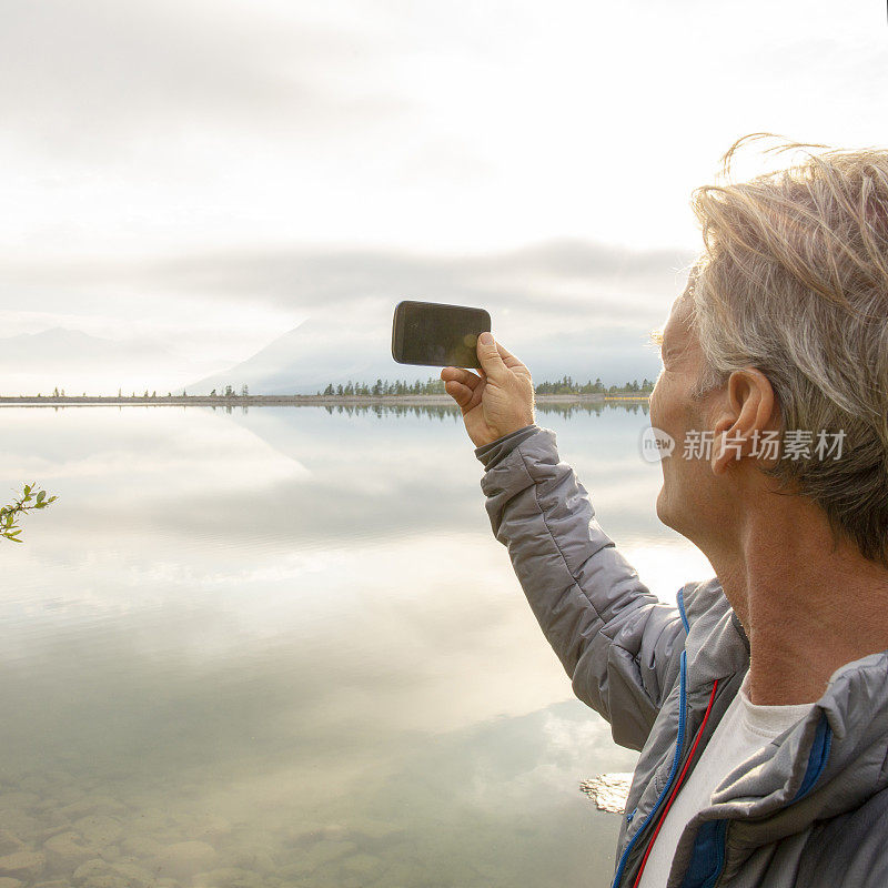 一名男性徒步旅行者到达湖岸，用智能手机拍下照片