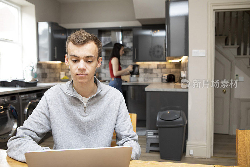 一名男大学生坐在厨房的桌子旁，用笔记本电脑工作，背景中有一名女学生正在泡茶