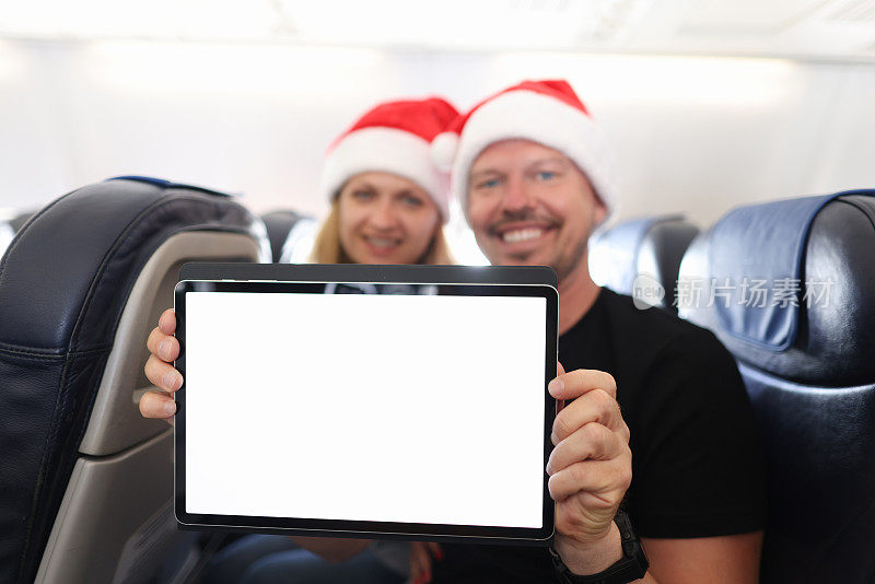一男一女戴着圣诞老人帽子，手里拿着电子平板电脑在机舱里