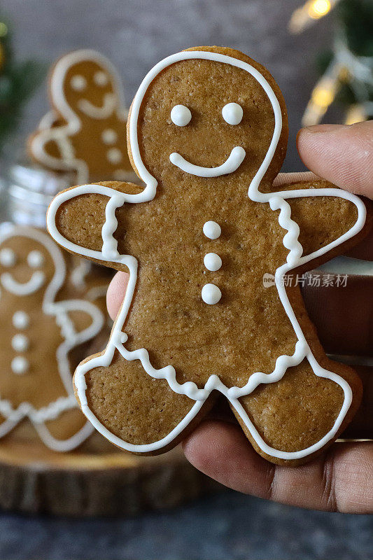 一个无法辨认的人拿着一个姜饼人饼干的特写图像，饼干上装饰着白色的皇家糖霜，姜饼人的玻璃饼干罐，迷你圣诞树，红色和金色的装饰物，灰色的背景，家庭烘焙