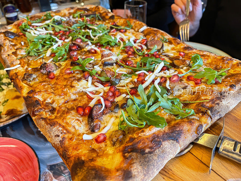 靠近一个巨大的方形披萨在桌子中央，吃饭时与朋友分享，有水果和蔬菜芝麻菜无花果和石榴，选择性聚焦