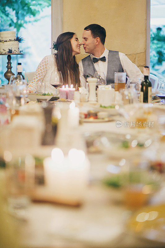 美丽的新婚夫妇庆祝婚礼的背景是美味的节日餐桌，在餐厅的食物和饮料。时尚快乐的新娘新郎亲吻。普罗旺斯婚礼