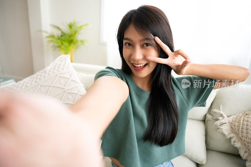 年轻漂亮的亚洲女性微笑着坐在客厅。她喜欢在模糊的背景下轻松愉快地自拍