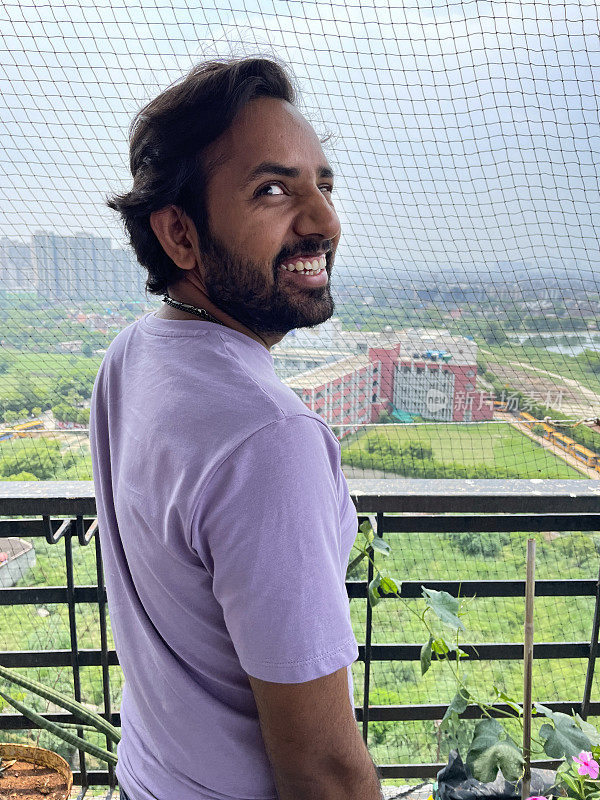 印度男子在公寓阳台户外的防虫网前摆姿势，微笑着看着镜头，城市景观背景，侧面视图，重点放在前景