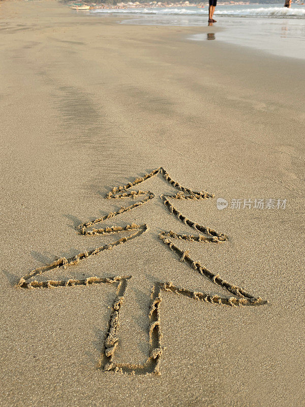 特写图片，圣诞树用木棍画在阳光明媚的水边沙滩上，背景是无法辨认的人在海里划水，重点放在前景，南半球的圣诞节概念