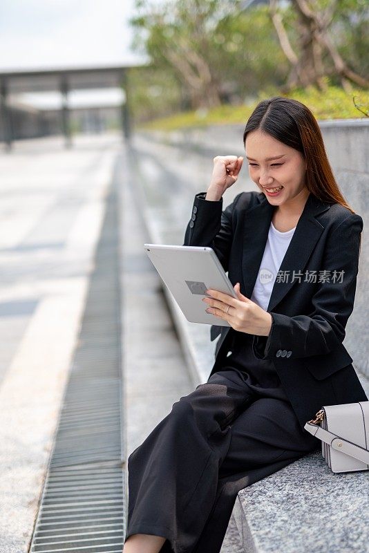 年轻美丽的亚洲女性办公室行政女员工显示yes获胜的姿态，在达到收入目标后，同时在她的办公大楼外的平板电脑上在线会见她的队友