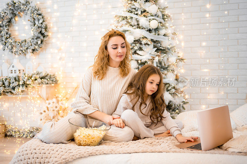 年轻女子带着7岁的小女孩穿着睡衣在圣诞装饰的家里看电影和吃爆米花
