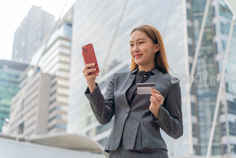 一名年轻的亚洲女商人穿着灰色的办公套装，在商业区用她的智能手机使用信用卡进行在线交易，她微笑着自信