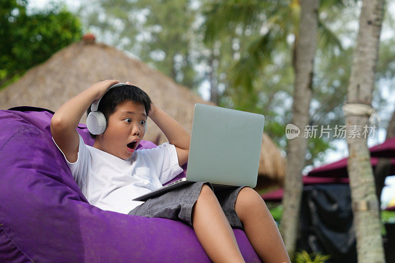 学校放假期间，一个可爱的亚洲男孩坐在一个豆袋上，惊讶地看着笔记本电脑。