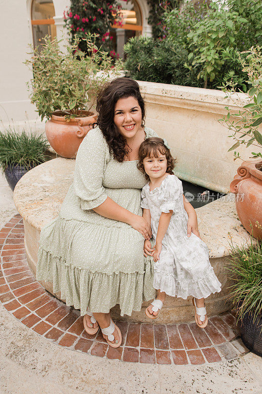 一个4岁的古巴裔美国女孩和她31岁的古巴母亲在佛罗里达州棕榈滩的托斯卡纳风格的花园里，穿着鼠尾草绿和白色的春装和白色的皮凉鞋，都是棕色卷发