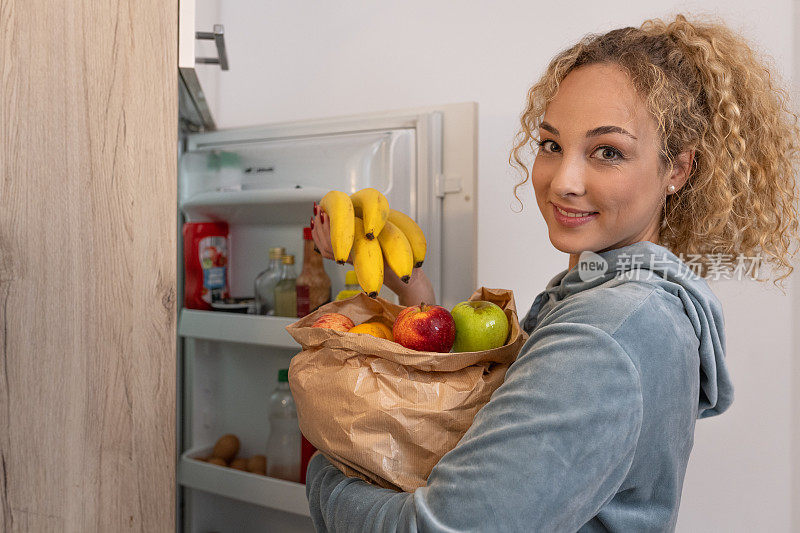 一个女孩站在冰箱前，拿着一个装满水果的购物袋