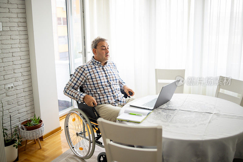 残疾白人中年男子坐在轮椅上，在家工作时用笔记本电脑进行视频通话