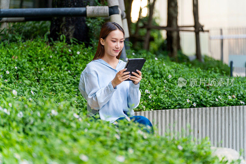 年轻的亚洲女性在办公大楼外的花园里使用平板电脑，喝着咖啡休息