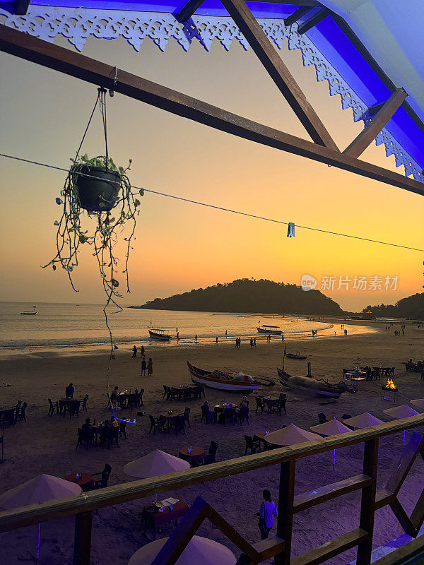 热带橙色日落天空的图像，Palolem海滩餐厅酒吧，果阿，印度，令人惊叹的日落在一排排的露天餐桌和遮阳伞上，从海滩小屋的阳台上观看