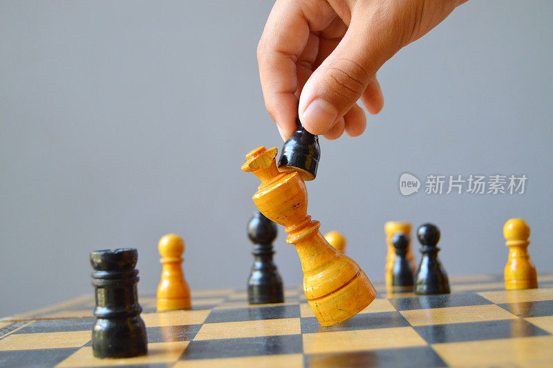 一只手拿着一个黑色的棋子，推或打白色的米色国王，使它摔倒或摔倒，在棋盘上被打败，描绘了失败的概念，损失，失败，挫折，在灰色的背景上有复制空间