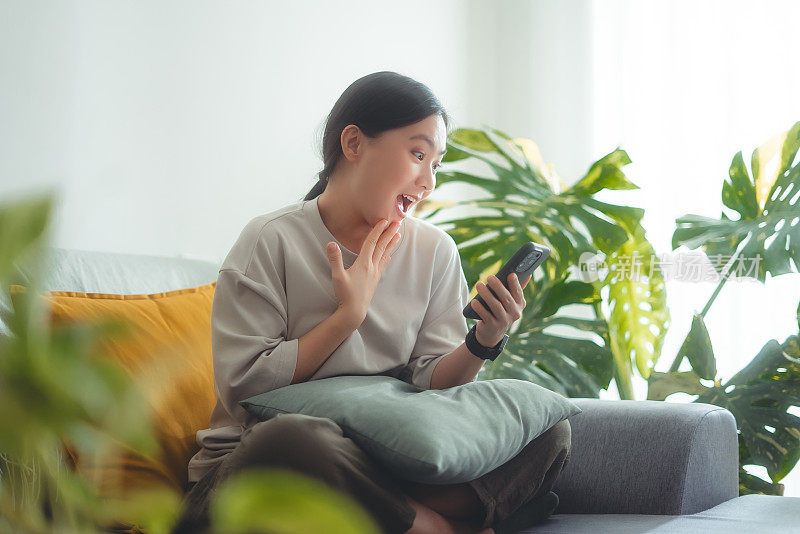 坐在家里客厅里玩智能手机的亚洲女人开心兴奋。