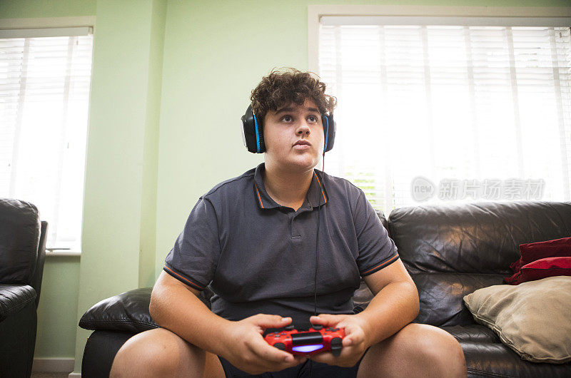 一个十几岁的男孩戴着耳机在玩电子游戏。他穿着灰色的t恤和短裤。