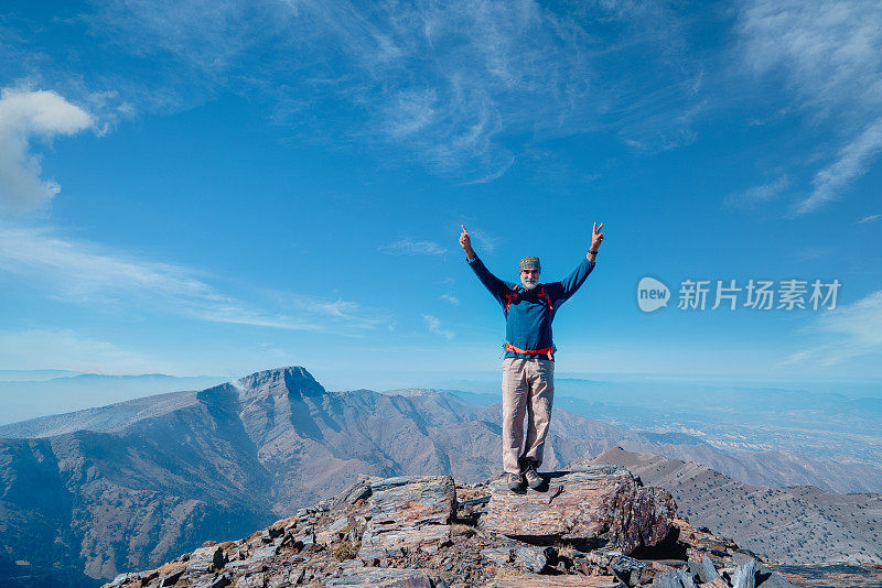 成功的年长登山者在山顶微笑着竖起大拇指，做着和平的象征