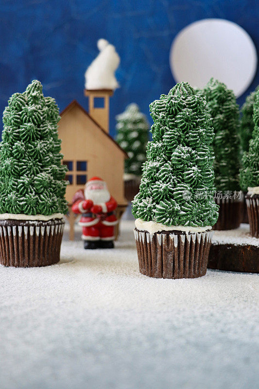 批量自制的形象，圣诞树设计的巧克力纸杯蛋糕在纸杯蛋糕盒展示在下雪，夜间针叶林场景，模型房子与圣诞老人，棉花糖雪人和满月，聚焦前景