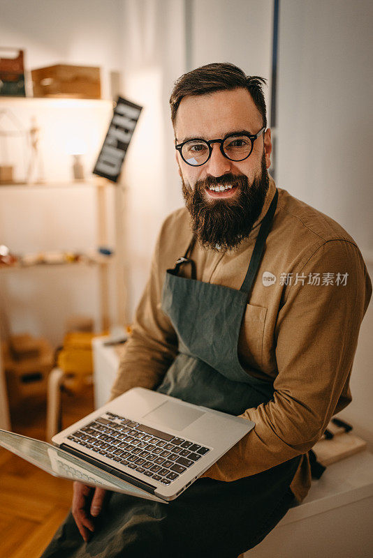 自信的大胡子艺术家的肖像与笔记本电脑坐在工作室
