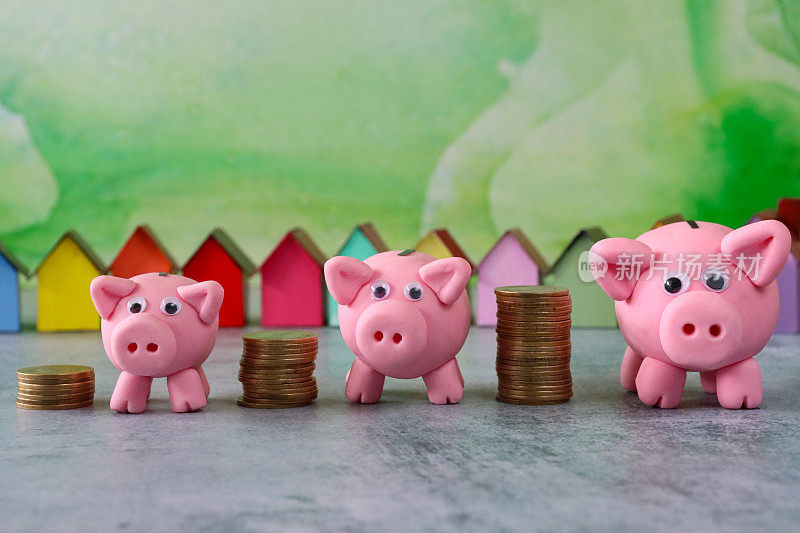 三个模型猪站在成堆的硬币前的一排排彩色纸板房子，彩色的海滩小屋，重点在前景，斑白的绿色背景，家庭财务，房地产和假日储蓄概念