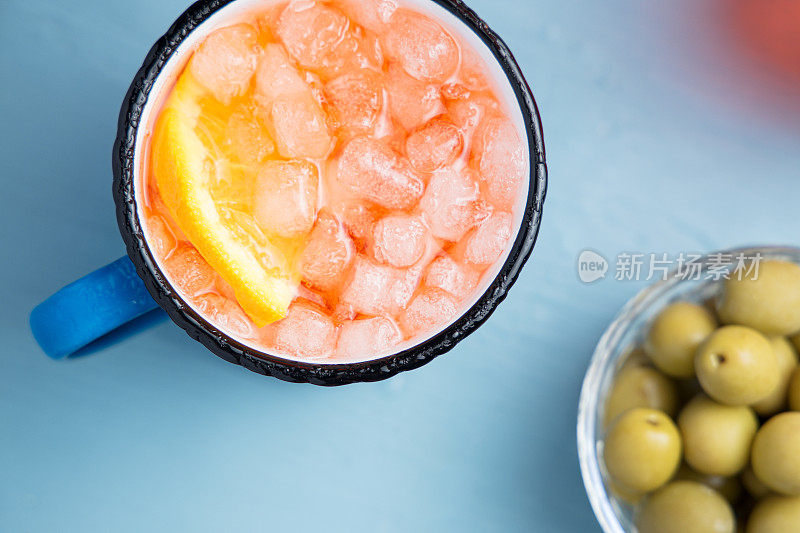 蓝色的桌子上放着搪瓷杯的经典意大利开胃酒喷淋鸡尾酒，配上一片橙子和橄榄。俯视图