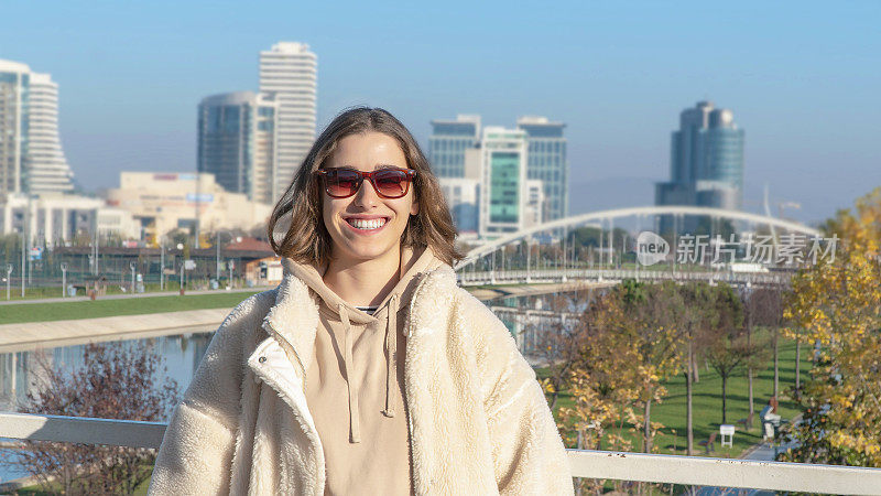 年轻女子快乐地站在户外微笑的肖像。年轻女子戴着太阳镜享受户外的新鲜空气。