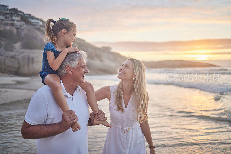 一个白人小女孩被她的爷爷抱着，而她的母亲在日落时走在海滩上的特写。一家人在夏日的阳光下玩耍
