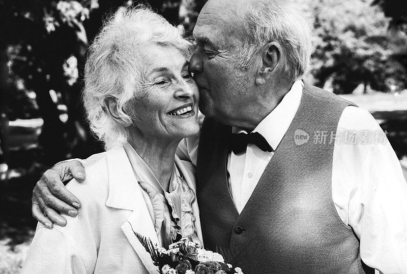 一对老夫妇在绿色的公园里散步。祖母和祖父在他们的金婚纪念日上。50年的爱情故事。奶奶和爷爷在接吻。黑色和白色。