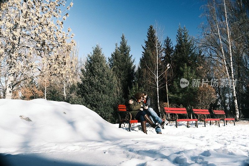 一对成熟的夫妇坐在冬天公园的长椅上，看着智能手机的屏幕