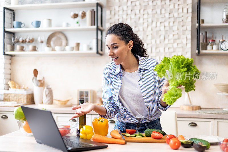 有用的烹饪和食物博客。积极的女人做新鲜的沙拉，看着笔记本电脑和手势，厨房内部