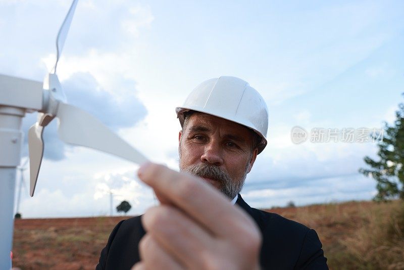 高级风力涡轮机能源工程师，穿着西装，戴着白色头盔，手持风车原型，在风力发电场，风力涡轮机，可再生能源从风。