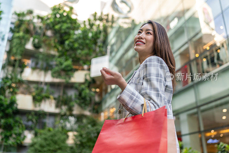 在一次成功的购物狂欢后，年轻的亚洲女商人在购物中心愉快地摆着购物袋，陶醉于购物疗法。