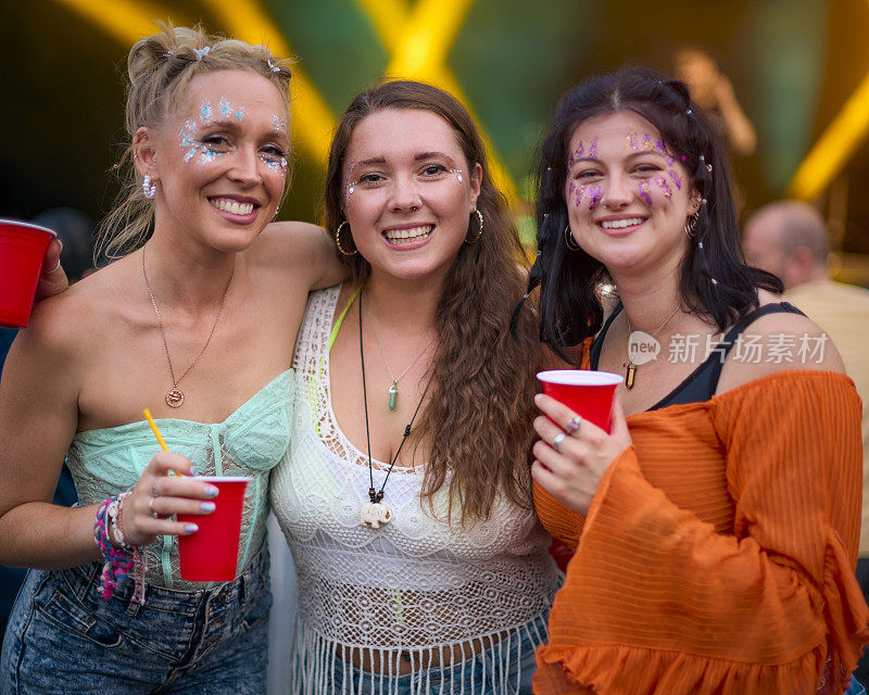 三位戴着亮片的女性朋友捧着饮料在夏日音乐节上玩乐的肖像