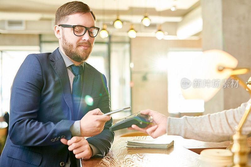 在餐馆付账时，穿着西装、满脸胡须、严肃自信的商人把智能手机装在电子阅读器上