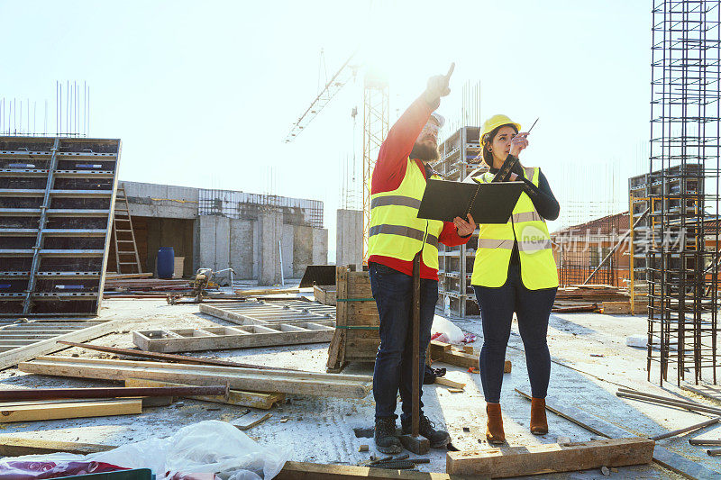 一个阳光明媚的日子，一位自信的女建筑师拿着平板电脑和一位建筑工人在一个住宅楼的工地上一起讨论和工作。