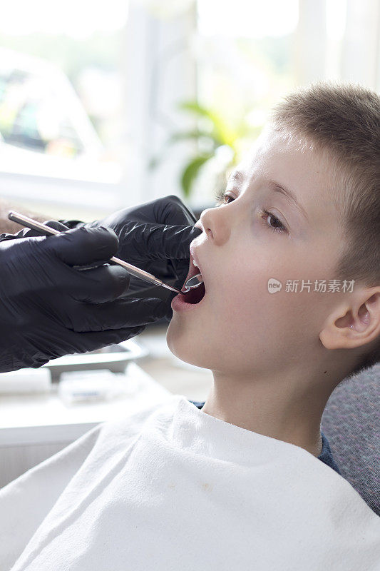 牙医的手上戴着一次性的黑色橡胶手套，拿着一面牙镜。这个学龄男孩张着嘴坐在椅子上。医生正在检查男孩的牙齿。