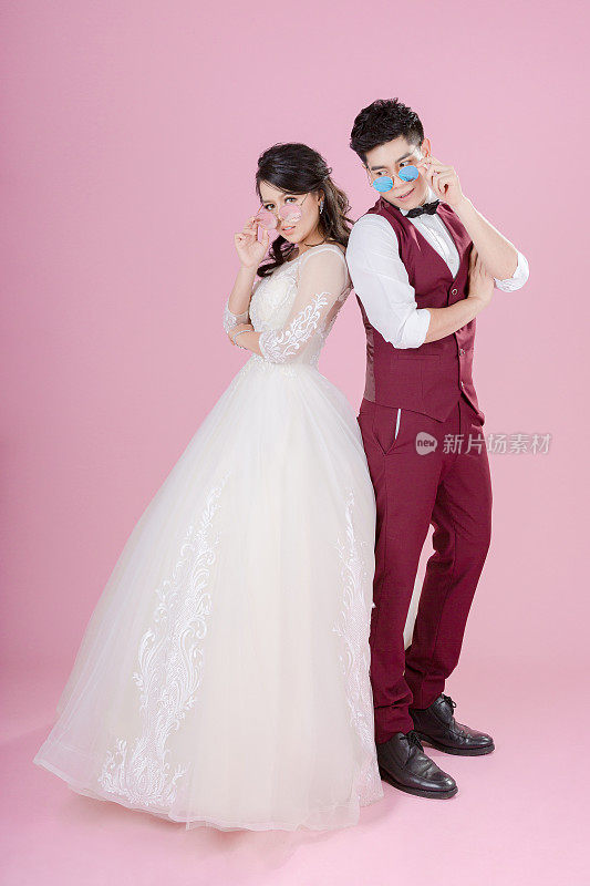 幸福的年轻新娘和新郎戴着太阳镜在粉红色的背景。新婚夫妇，新家庭，婚纱。新娘的婚礼。爱的概念