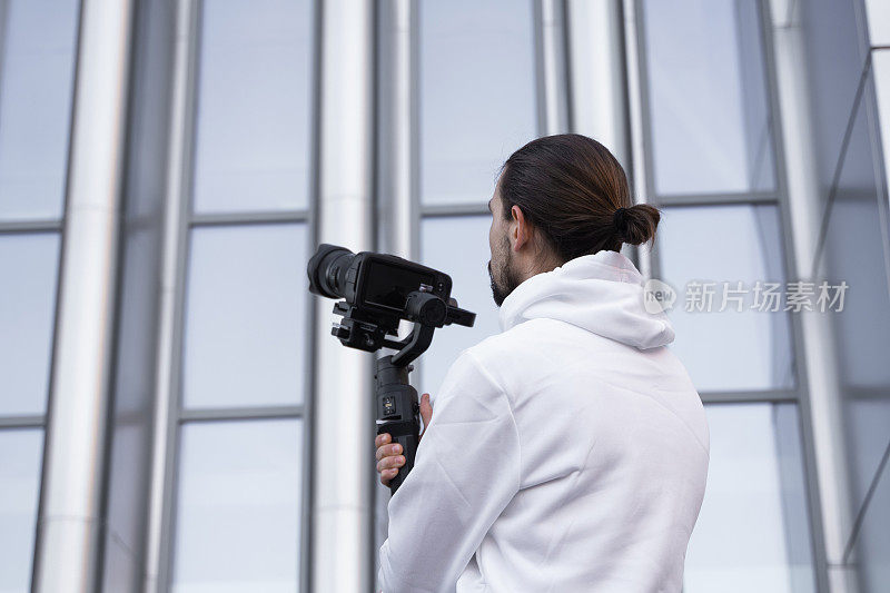 年轻的专业摄像师手持专业摄像机，安装在三轴框架稳定器上。专业设备有助于制作高质量的视频而不摇晃。一个穿着白色连帽衫的摄影师正在录像。