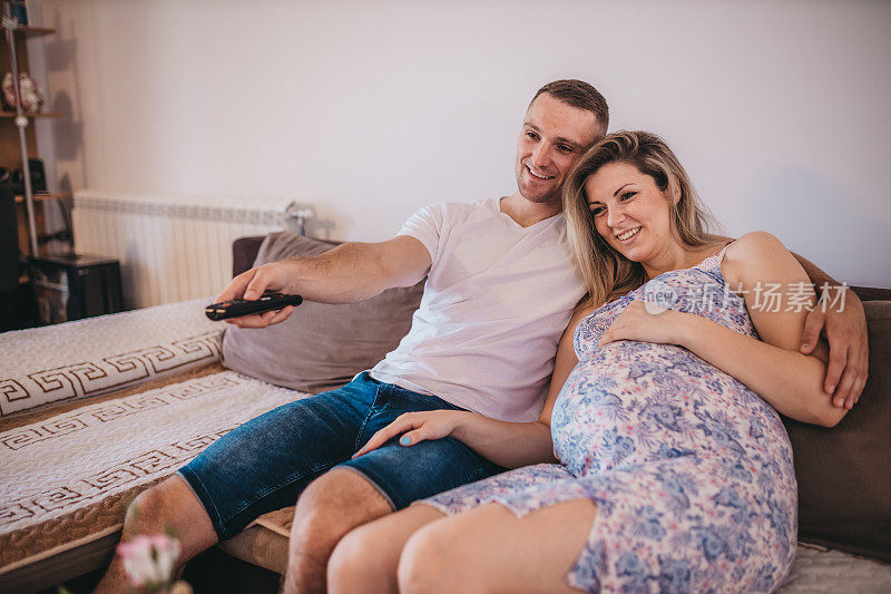 年轻的丈夫和他怀孕的妻子坐在家里的沙发上看电视