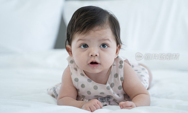 3个月大的新生婴儿躺在白色的床上。天真可爱的混血婴儿，亚洲人和高加索人，正在撒谎或爬行。小女孩在家里穿着可爱的衣服。本空间
