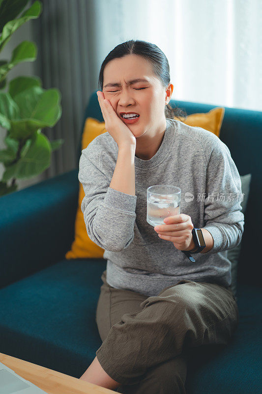 亚洲女性坐在客厅的沙发上，喝了凉水，摸了摸脸颊，就会感到牙齿敏感。