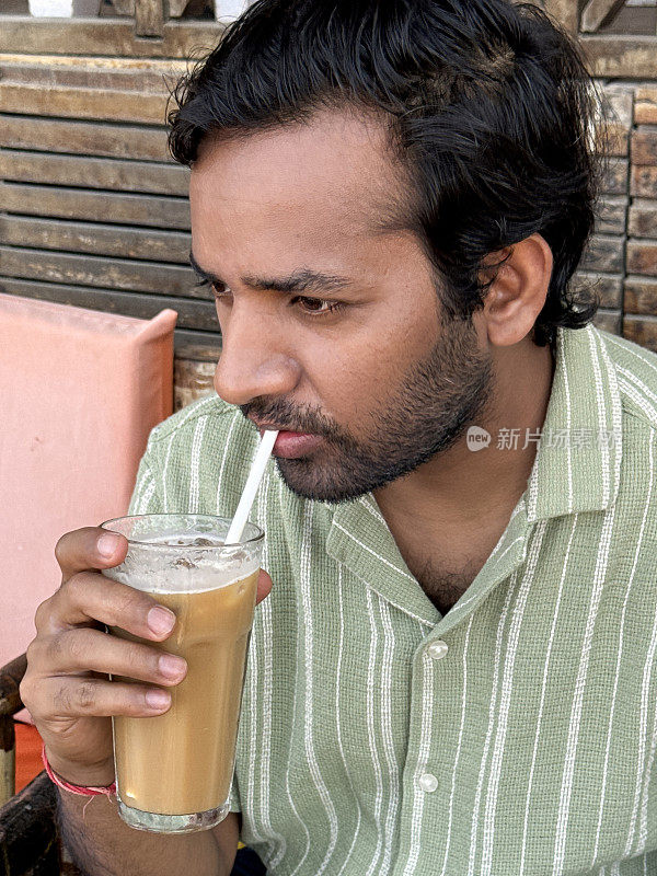 印度人坐在餐厅的露天用餐区，户外扶手椅，拿着一杯冰咖啡饮料，纸吸管，木板背景的面板墙，重点在前景