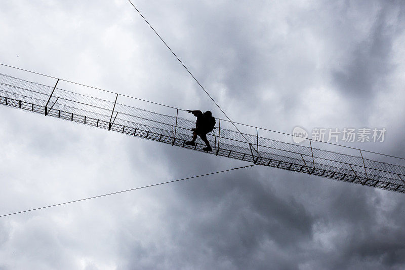 一名年轻女子在高空穿过一座悬索桥，背景是乌云