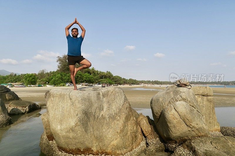 印度人在岩石上练习树姿势(Vrksasana)瑜伽姿势，水的边缘，帕洛伦海滩，果阿，印度