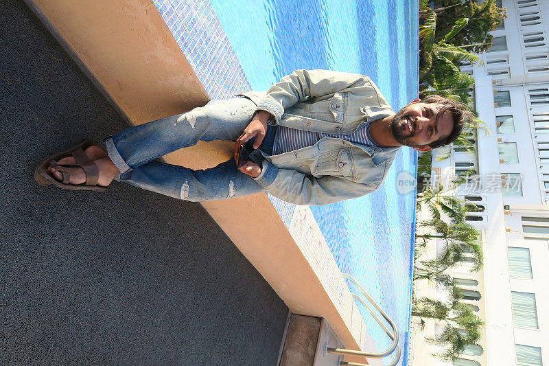 英俊的年轻印度男子坐在游泳池在阳光下看起来快乐的暑假，牙齿微笑，大笑，手在头发，好看的印度男子享受阳光无限游泳池蓝色马赛克瓷砖，度假模式在牛仔夹克