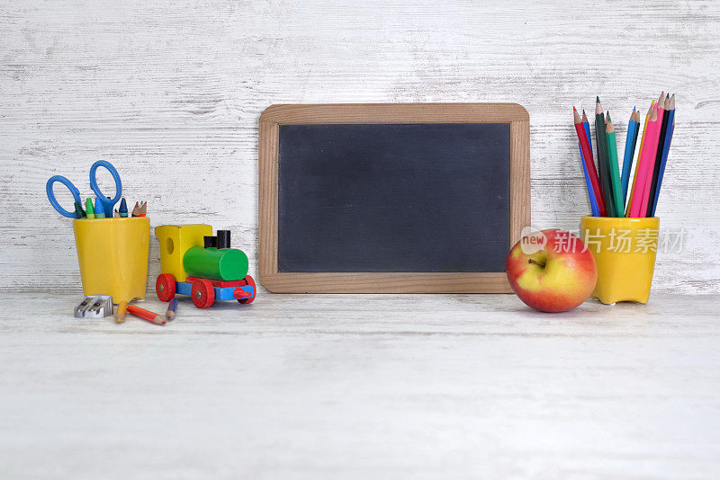 儿童书桌上有一块黑色的黑板，黄色的罐子里有几支彩色的铅笔和一个苹果
