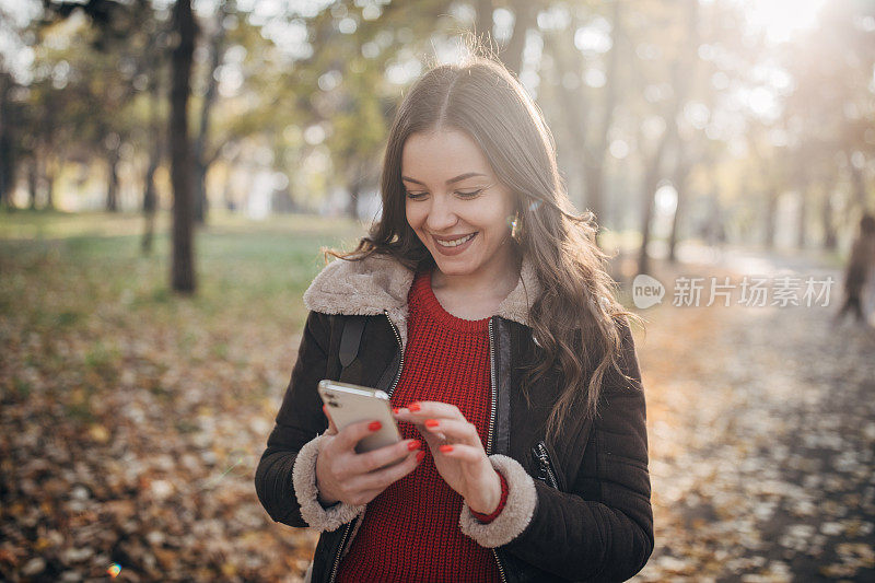 快乐的女人在秋天的公园里散步，用着智能手机
