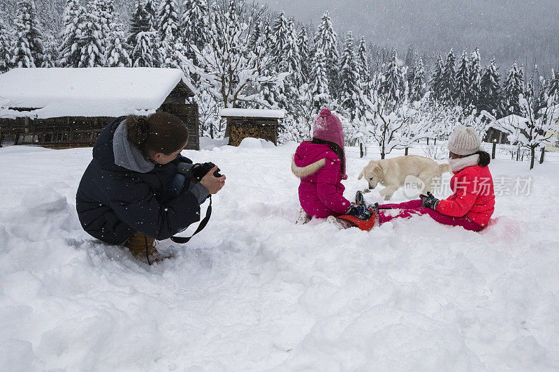 当女孩们和狗在雪中玩耍时，妈妈给他们拍了照片