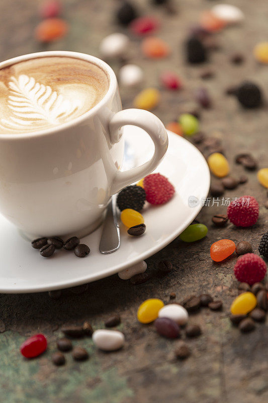 热咖啡拿铁杯与罗塞塔拿铁艺术牛奶泡沫，咖啡豆和彩色果冻糖果在质朴的木头桌子背景